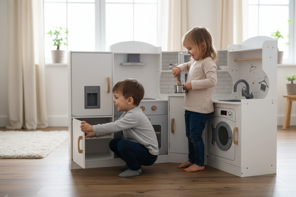 Two children playing with a white toy kitchen set in a bright room with wood floors and large windows. One child is crouching and opening a cabinet door, while the other stands beside the stove and sink area.