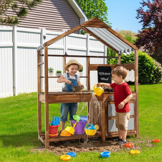 A children's wooden play kitchen set in a backyard, with two kids playing inside and around the structure. The wooden play kitchen features a house-shaped frame, a gray-and-white striped awning, a small black sign reading 'Kitchen Mud,' and multiple colorful toy buckets, shovels, and cups placed underneath and on the grass.