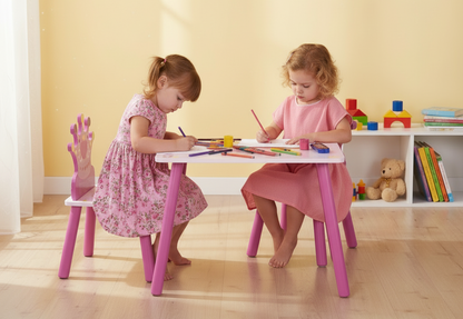 Two young girls sitting at a pink table and chairs set, drawing with colorful crayons in a bright room.