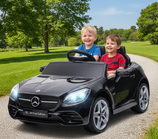 Two children in a black Mercedes-Benz toy car on a road with greenery in the background